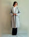 Model holding the lapels of the open grey SoHo Belted Overcoat, showing its long length and side slits