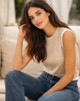 Brunette model in a sand sleeveless knit top styled with dark jeans, seated casually on a sunlit patio.