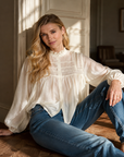 Ivory Sheer Poet Blouse with ruffled stand collar and balloon sleeves, worn with blue jeans while seated on a wood floor