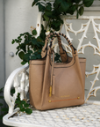 Montemare Sedona Crossbody in Latte styled on a white garden bench, showcasing its braided handles, structured silhouette and gold hardware.