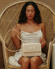 Model seated in a rattan chair holding the Oceans End Giovanni crossbody in creme