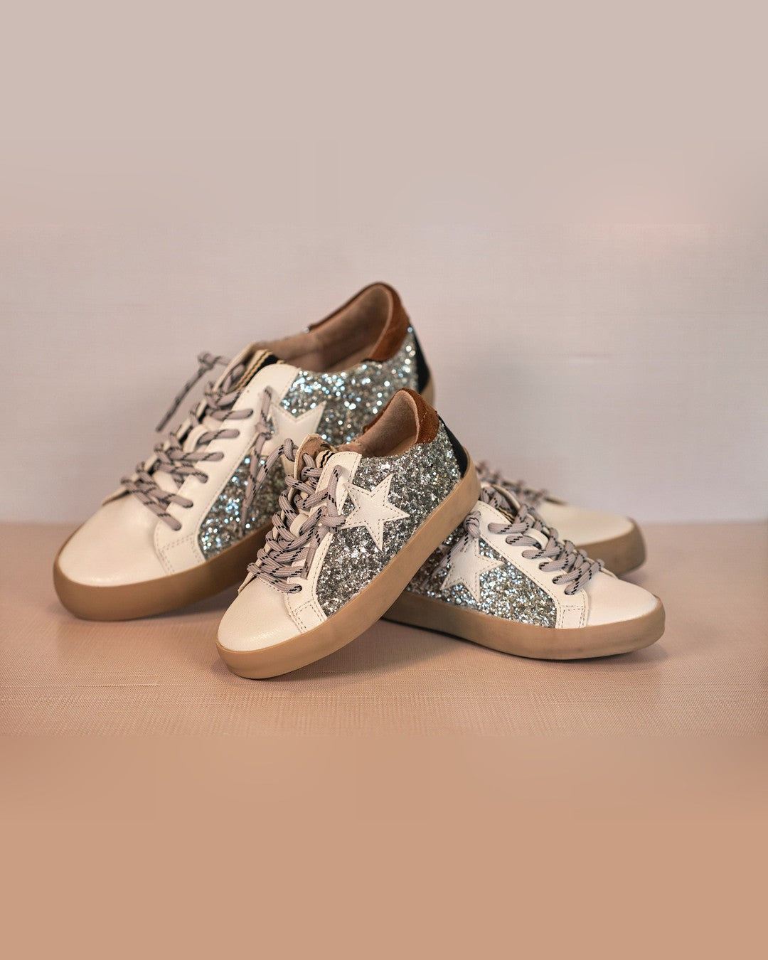 Two pairs of Shushop Paula Glitter Sneakers stacked together on a wood surface