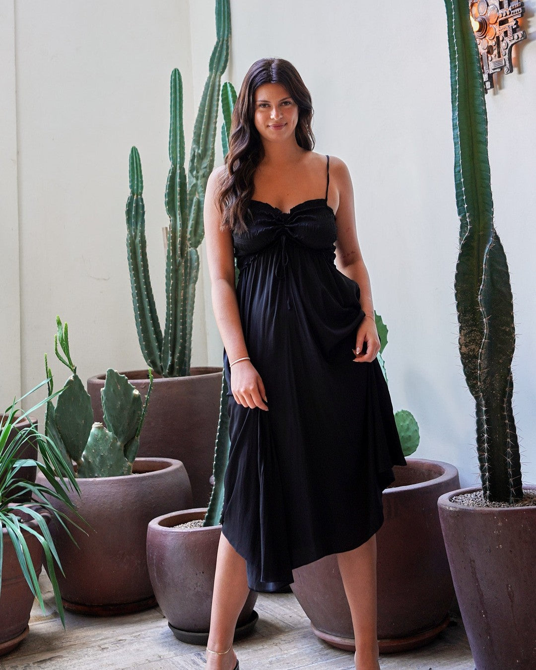 Model in the black Prescott Midi Dress with tie bust and black heeled sandals, standing among potted cacti