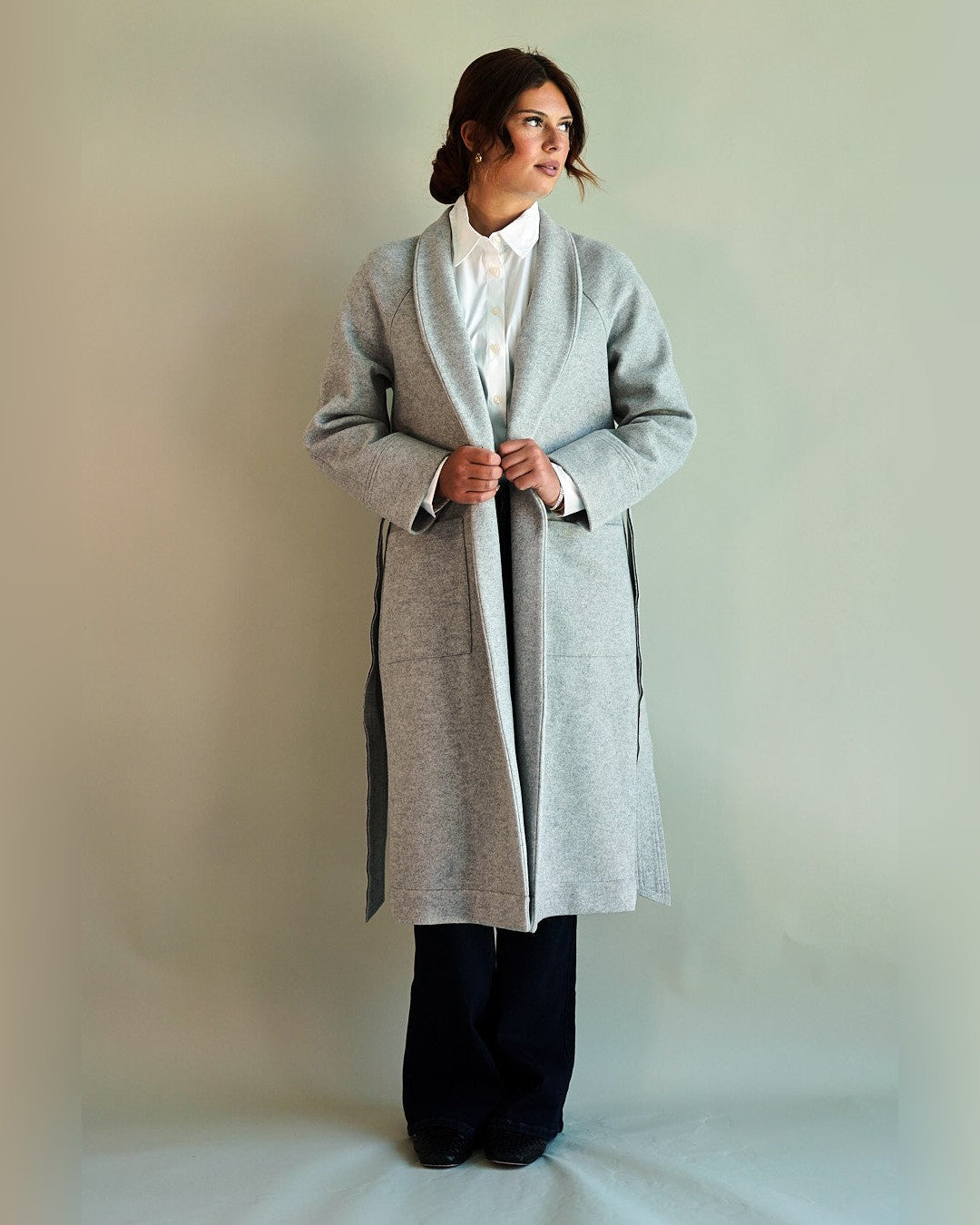 Model holding the lapels of the open grey SoHo Belted Overcoat, showing its long length and side slits