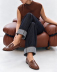 Model in a brown knit and cuffed jeans seated on a leather chair wearing Dolce Vita Baxen russet loafers