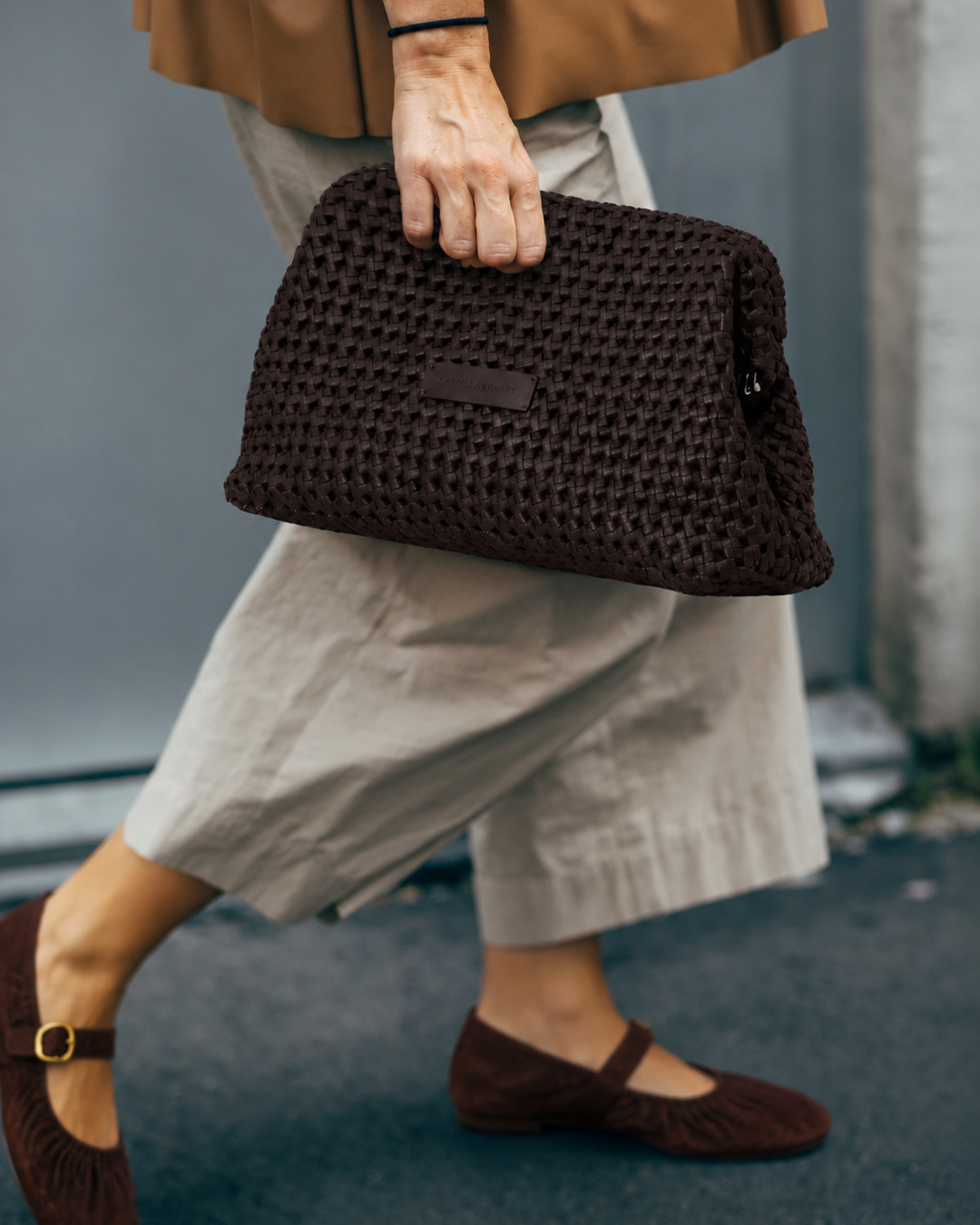 Model in cream trousers and burgundy Mary Janes carrying the Daniella Lehavi Vivian Maxi Clutch in chocolate by hand
