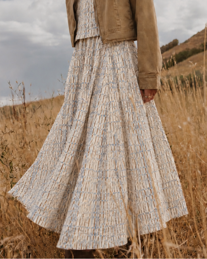 A woman stands in a dry grassy field under a cloudy sky, wearing a long cream patterned maxi dress with a tan suede-look jacket.