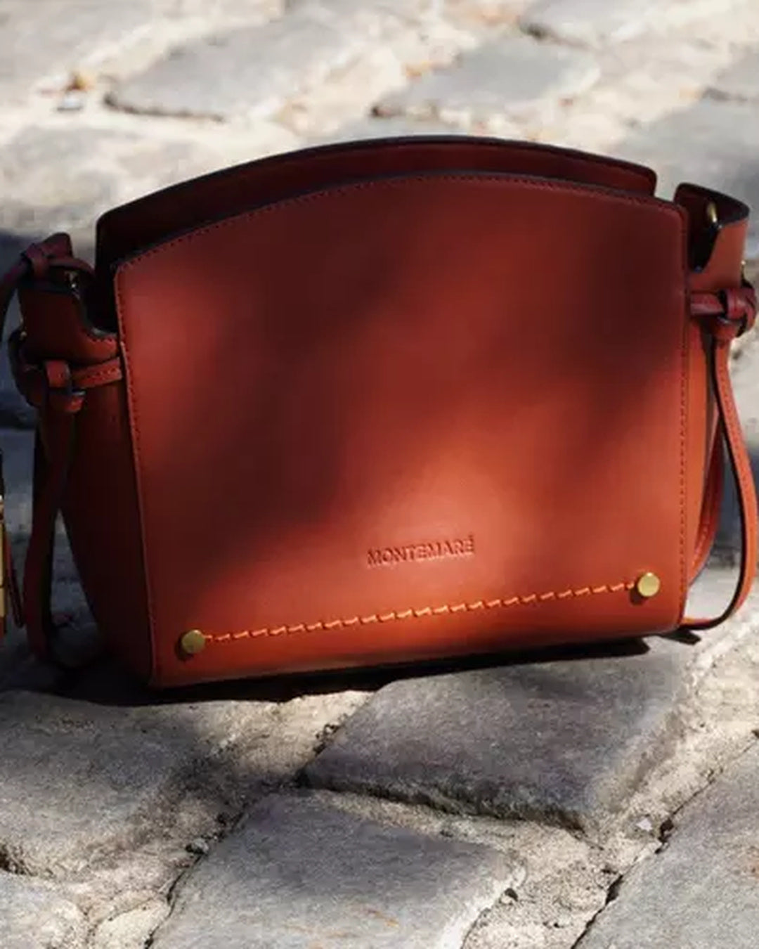 Montemare Roma Crossbody Bag in terracotta displayed on cobblestone with gold-tone hardware and contrast stitching