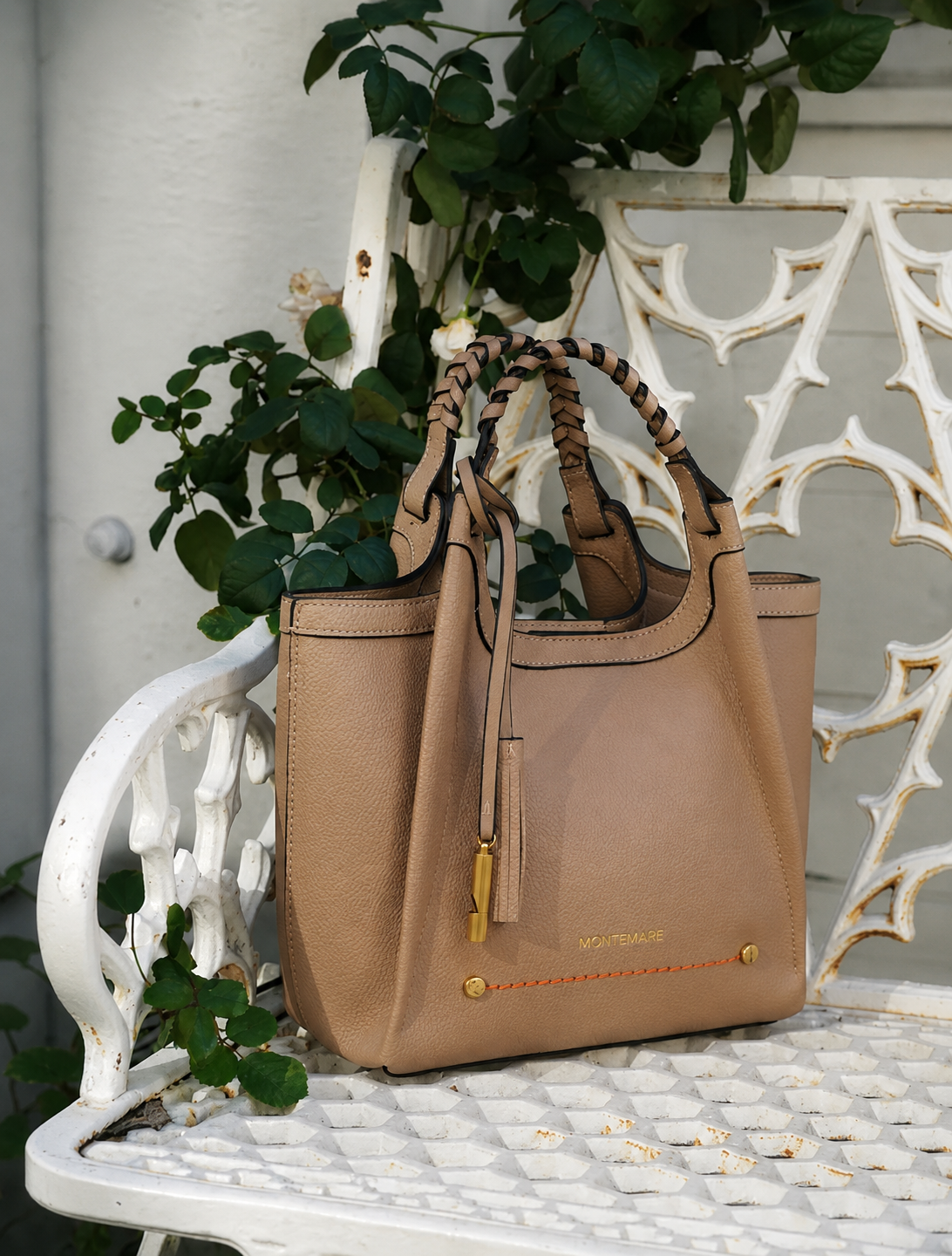 Montemare Sedona Crossbody in Latte styled on a white garden bench, showcasing its braided handles, structured silhouette and gold hardware.